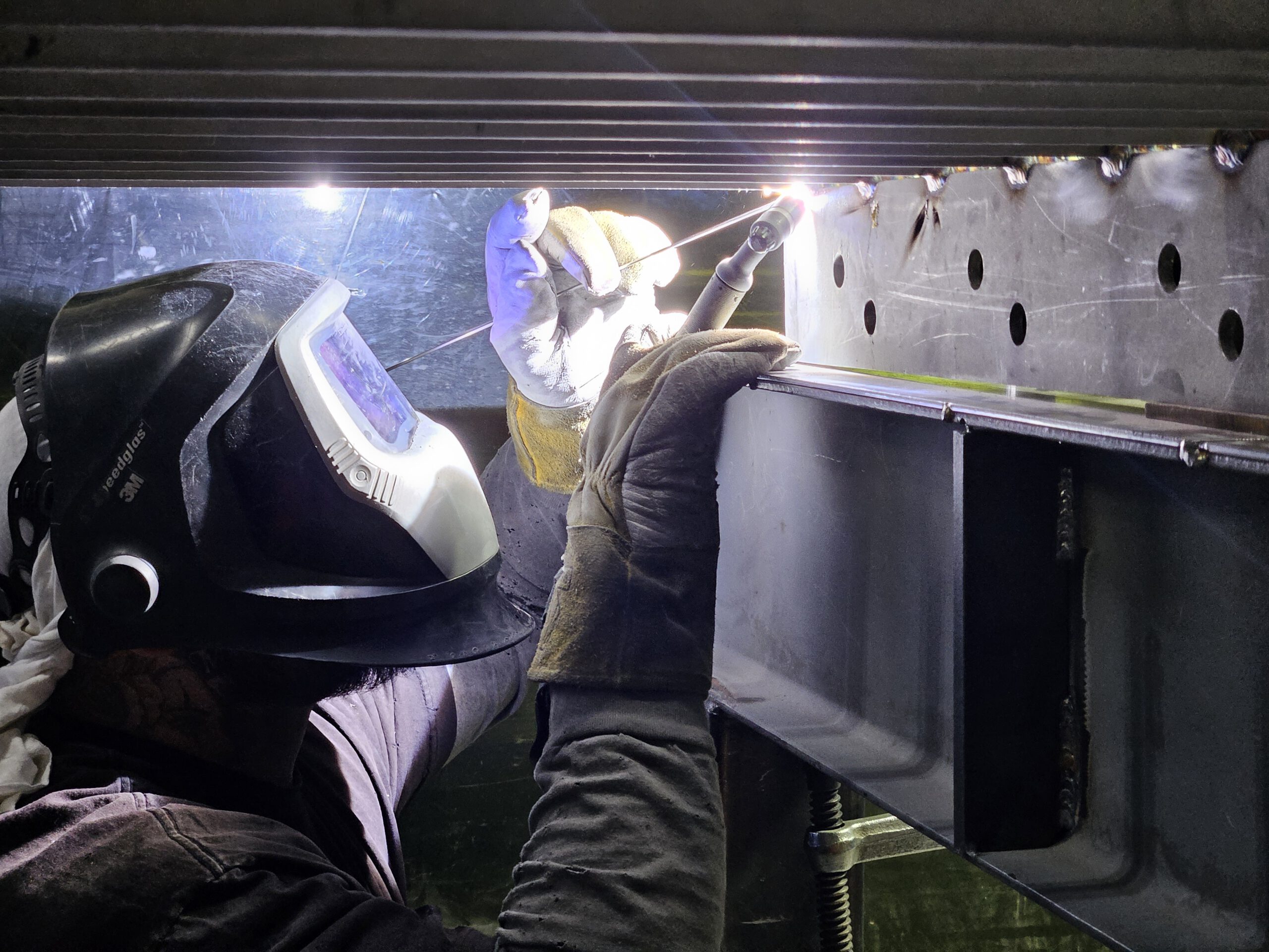 Person_With_Protective_Mask_And_Gloves_Welding_Underneath_Metal_Structure